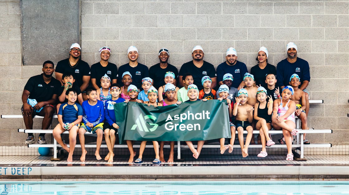 A group of children and adults pose for a photo, holding a banner that reads Asphalt Green.