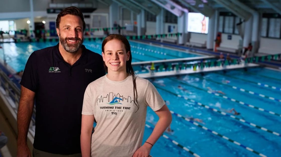 Senior Director of Aquatics, David Rodriguez, and swimmer Ali Wright smiling to camera with Asphalt Green's Upper East Side pools in the background.