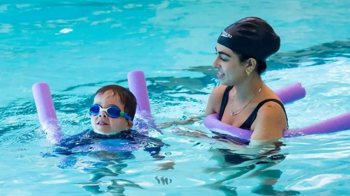 A child and an adult in the pool holding pool noodles.