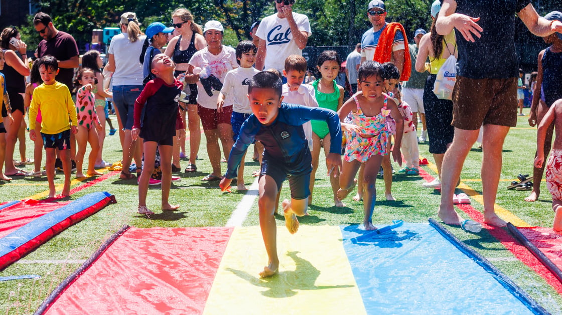 Children run down a slip-n-slide on Sprinkler Day at Asphalt Green.