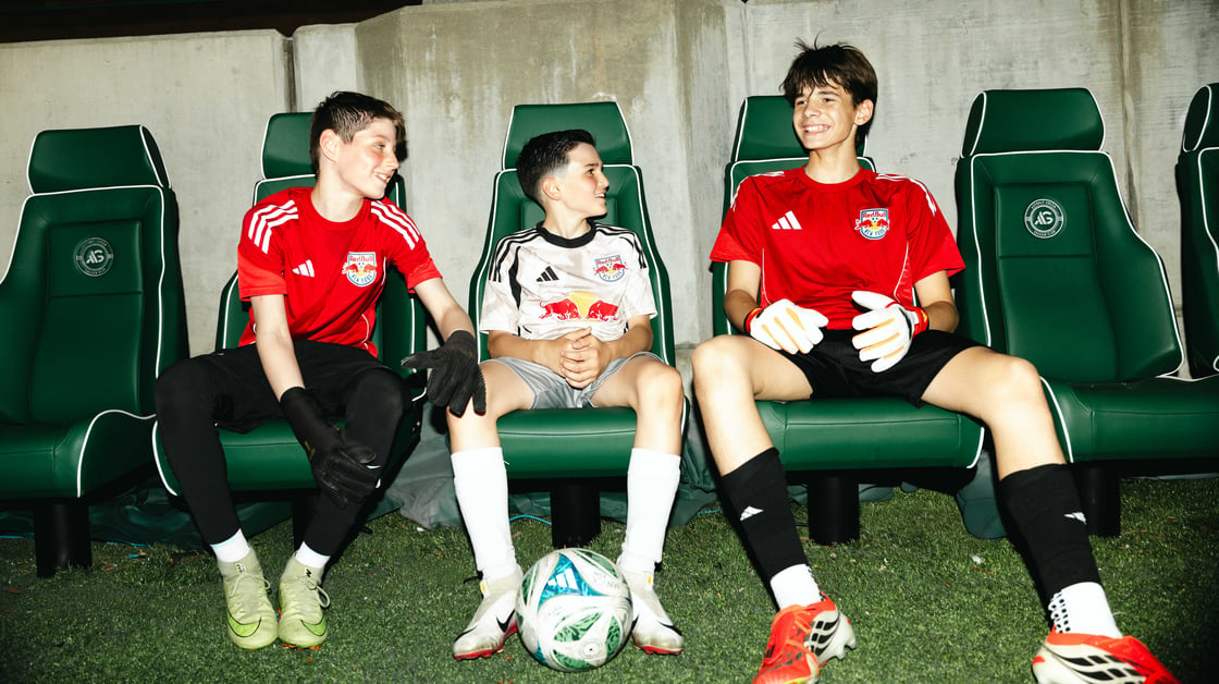 Three AGSC players smiling in uniform sitting on a bench with a soccer ball between them.