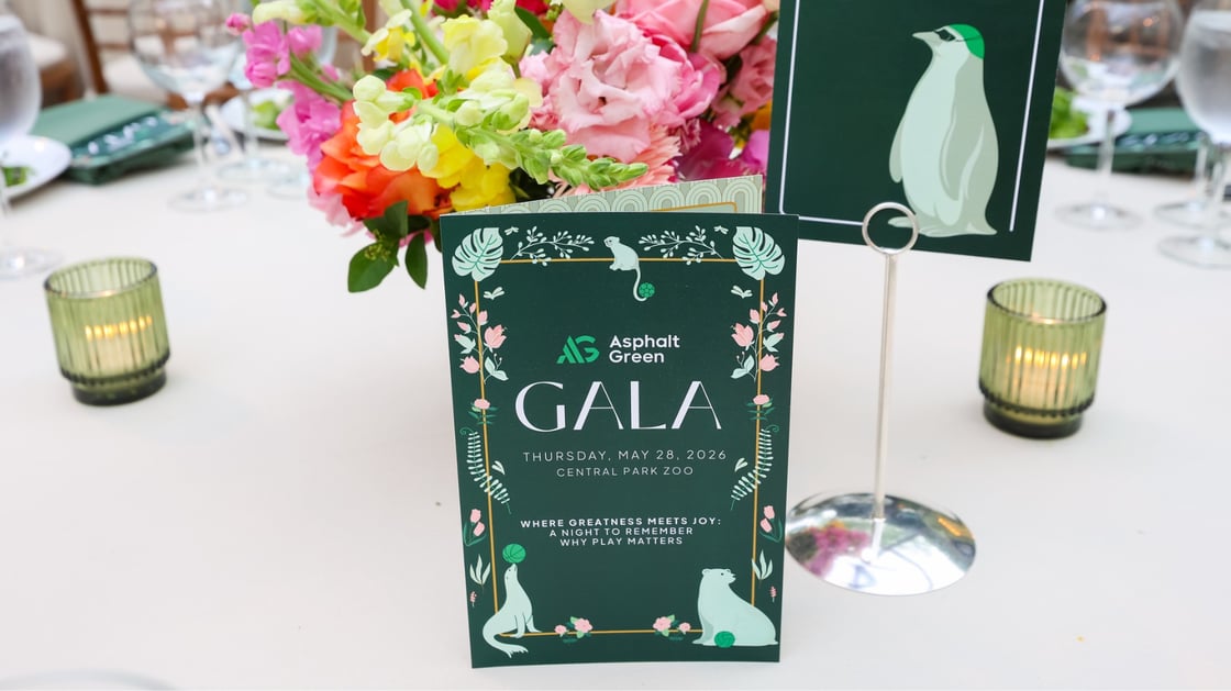An Asphalt Green Gala 2026 program card in front of flowers and candles on a table.